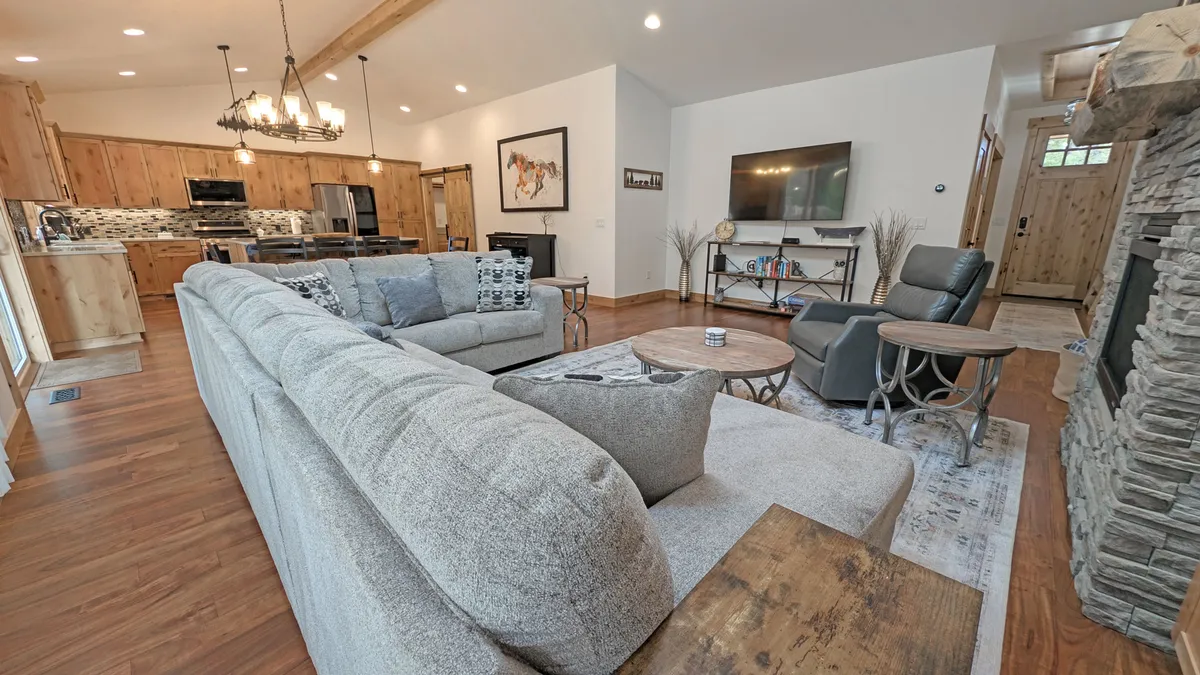 The hero image showcases a spacious, sunlit open-concept living area featuring a plush sectional sofa, elegant wooden dining set, a striking stone fireplace, and stylish wall art. The inviting space is designed for gatherings, with plenty of room for relaxing, sharing meals, and enjoying cozy evenings by the fire.