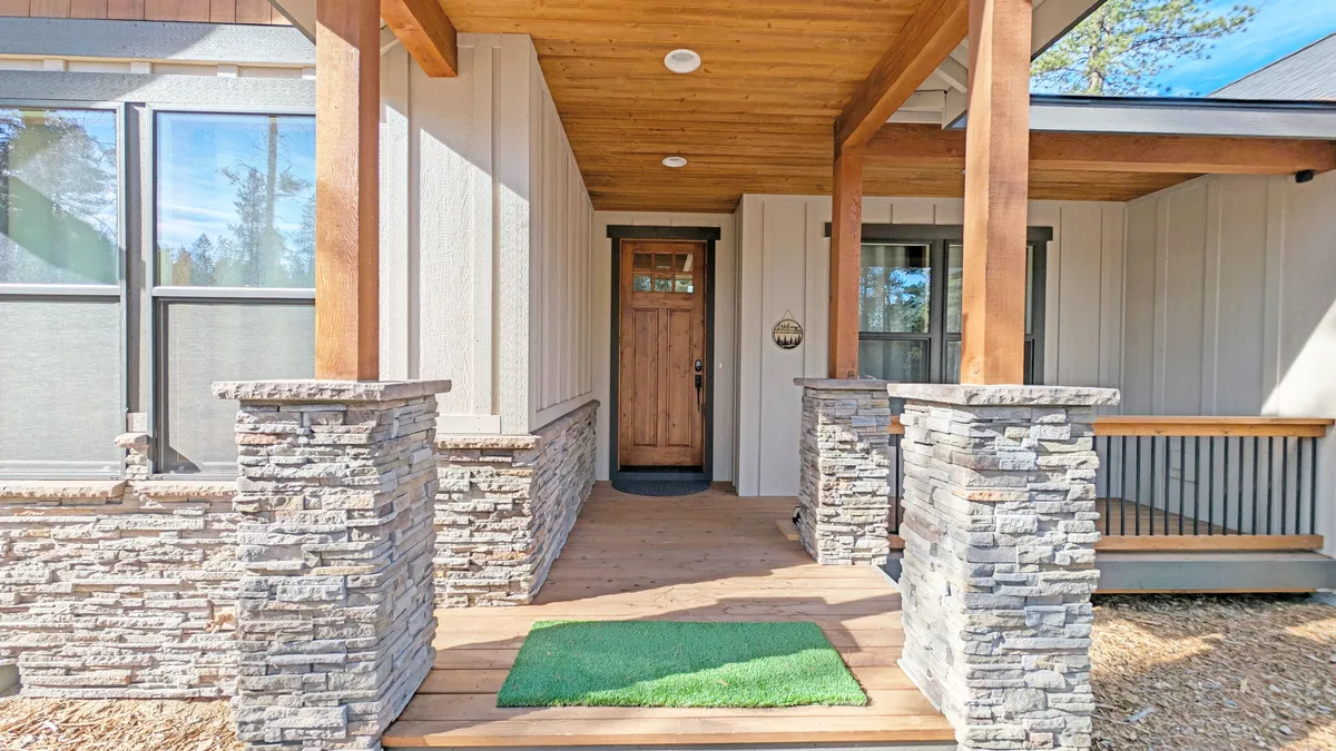 This exterior shot features the front door framed by striking stone columns, a wooden ceiling, and an inviting green mat. The entryway exudes a sense of welcome and hints at the tranquility found just beyond the threshold, while the outdoor materials echo the natural surroundings of Sunriver.