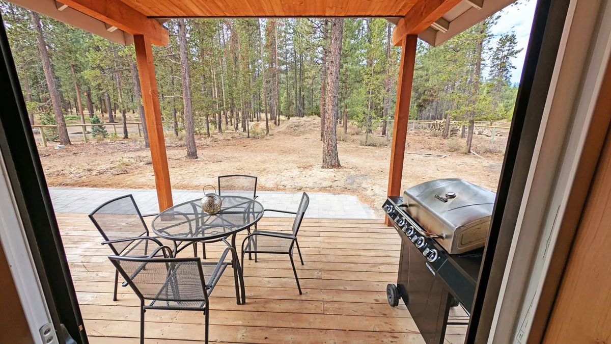 An outdoor dining area nestled among tall trees, with a table and chairs inviting guests to gather, and a grill ready for barbecues. The tranquil setting showcases the natural environment and opportunities for al fresco meals or relaxed evenings after exploring the Village.
