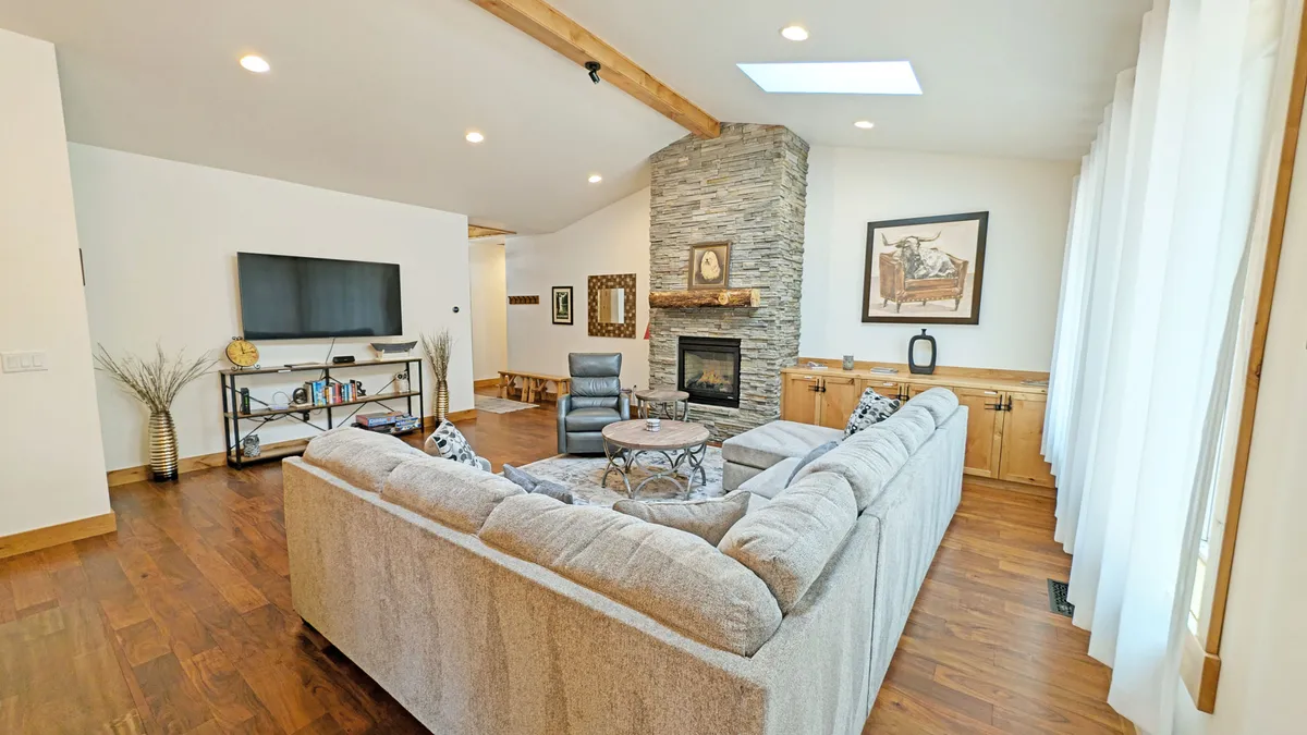 A cozy, open-concept living room with a large sectional sofa, stone fireplace, TV unit, and expansive windows letting in natural light. The space is arranged for movie nights, conversation, and relaxing with loved ones.