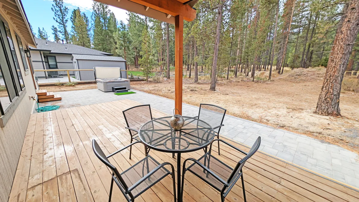 An inviting outdoor deck features a glass table with chairs surrounded by trees, perfect for outdoor dining, social gatherings, or relaxing after a day of exploring lakes and mountains. The tranquility and connection to nature is palpable.