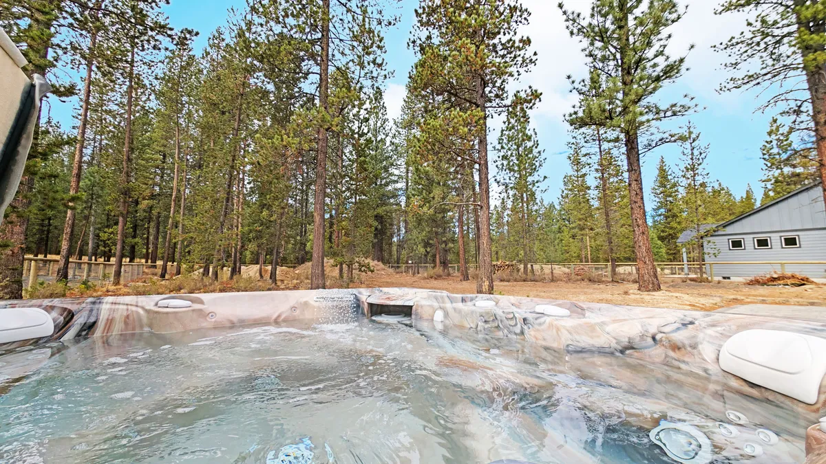 A relaxing hot tub sits nestled among tall pine trees, with a view of a distant cabin in the background. The setting communicates a peaceful retreat, surrounded by nature’s tranquility.