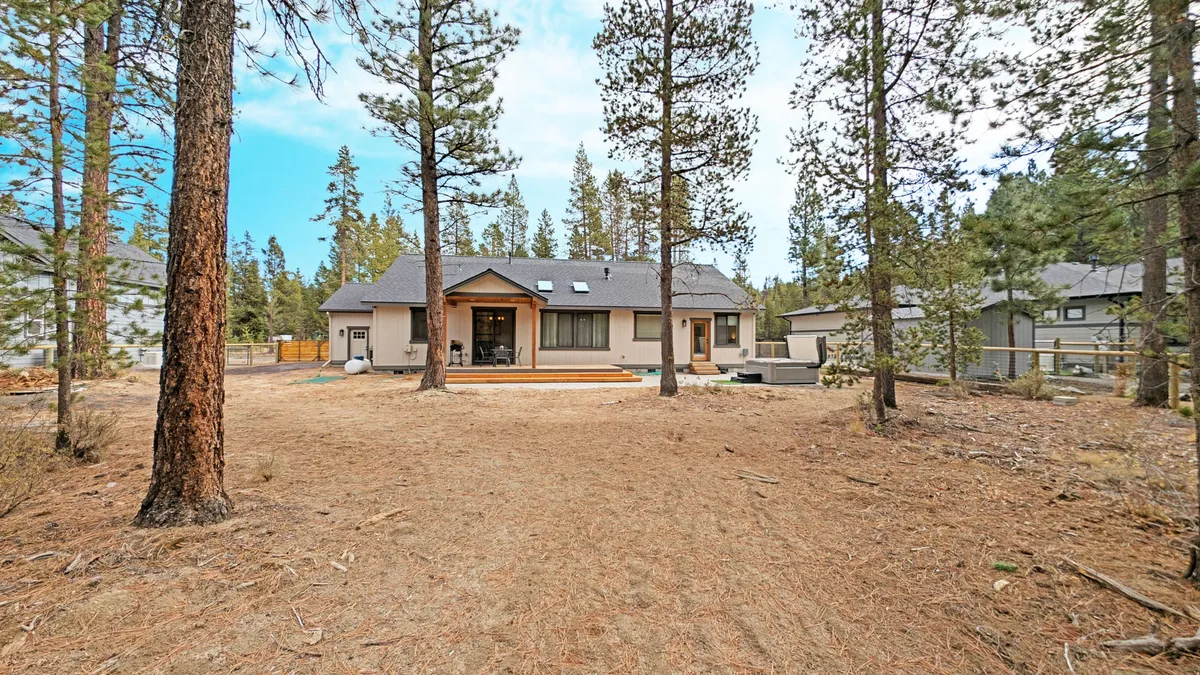 This image presents the home's exterior with the paver deck extending into the backyard, bordered by towering pine trees. The deck is spacious, inviting outdoor gatherings or relaxing moments. The natural forest setting is prominent, capturing the essence of privacy and forest access.