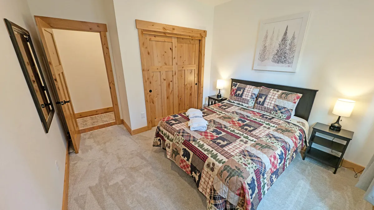 A comfortable, light-filled bedroom with a quilted bed, wooden sliding closet doors, nightstands with lamps, and framed artwork. The room feels peaceful and homey, ideal for couples or grandparents.