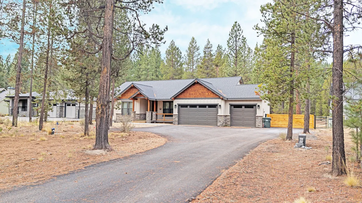 The exterior of the property displays modern architecture with welcoming wooden accents and large garage doors, sitting comfortably within a landscape of trees and natural elements. The image conveys both curb appeal and the connection to the greater Sunriver setting.