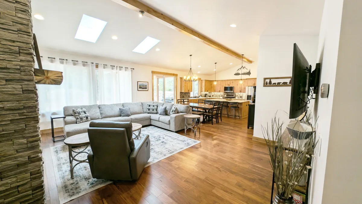 This image captures the open-concept main living area, with a spacious sectional sofa, a butterfly-style dining table, and a modern kitchen all visible. The space is accented by natural wooden beams and skylights, allowing plenty of natural light to infuse the room. The warm glow from the gas fireplace creates an inviting atmosphere, and the kitchen features a large island and gleaming stainless steel appliances.