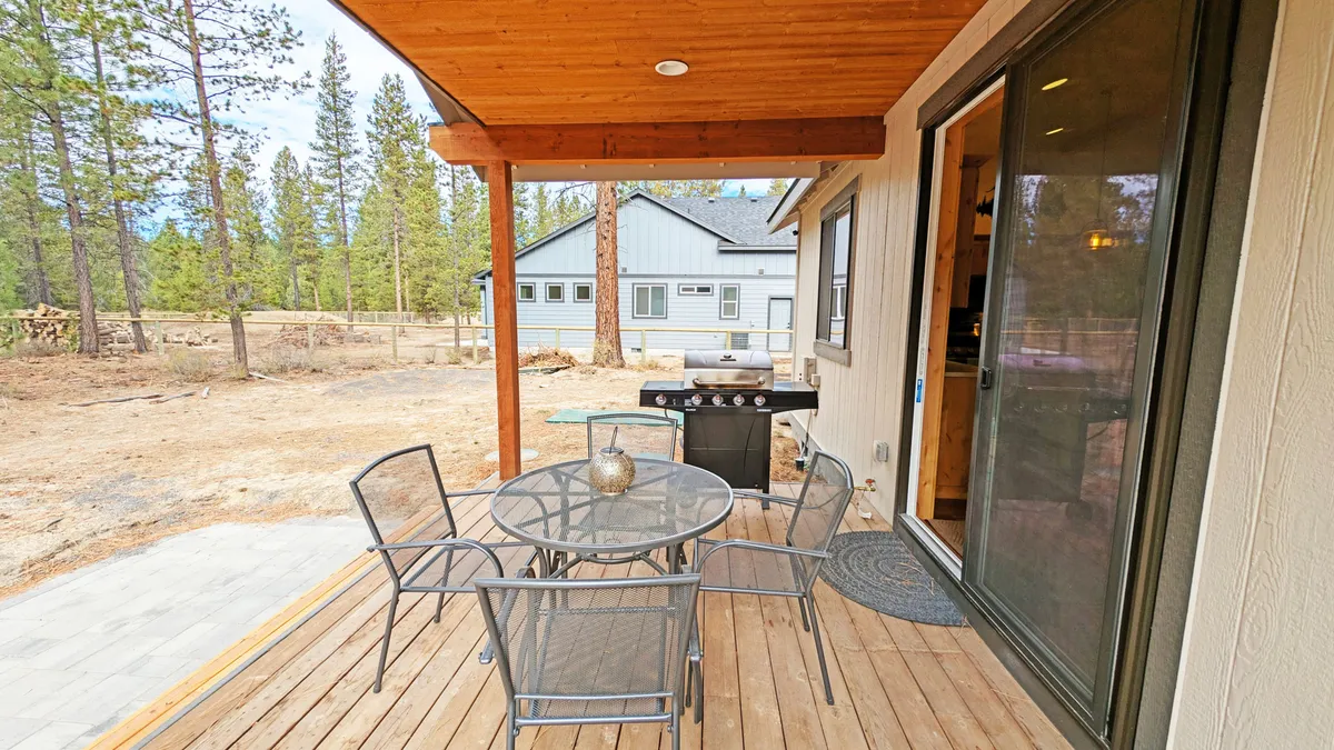 A spacious wooden deck featuring an outdoor dining table and grill, nestled among the trees. The setup is perfect for alfresco meals, barbecues, and gathering with friends or family while enjoying the natural surroundings.