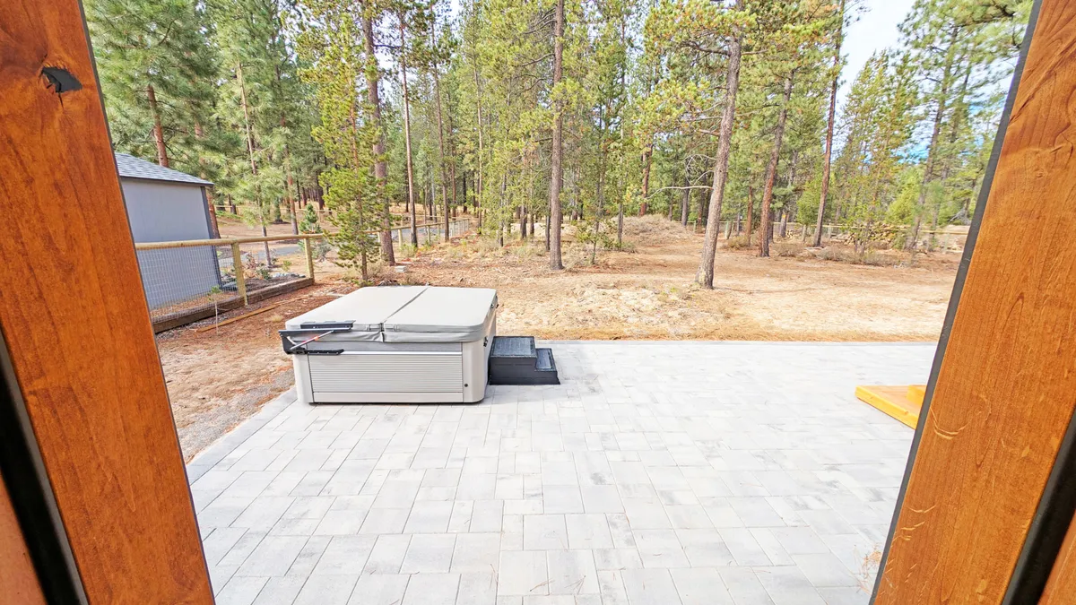This image features the hot tub nestled on a patio area, surrounded by tall pines. The ambiance is serene, with the hot tub positioned under cover, allowing guests to enjoy a soak regardless of the weather. The patio is adjacent to nature, highlighting the close proximity to the forest.